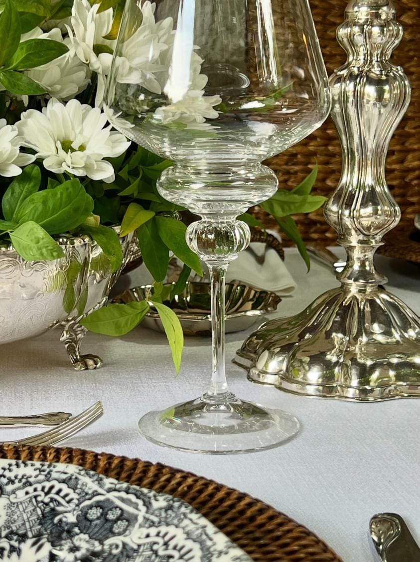 Copa entre cubiertos y elementos de servicio, con flores en el fondo.